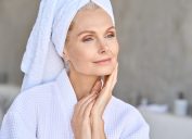 Happy beautiful middle aged woman wearing bathrobe and white towel with perfect complexion touching face looking away in bathroom. Advertising of skin care spa wellness concept. Closeup portrait.
