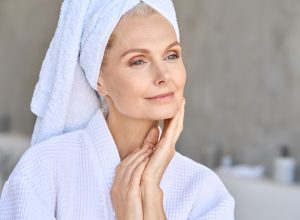 Happy beautiful middle aged woman wearing bathrobe and white towel with perfect complexion touching face looking away in bathroom. Advertising of skin care spa wellness concept. Closeup portrait.