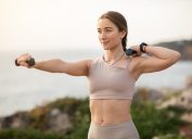 Glad confident strong millennial caucasian woman athlete in sportswear doing arm exercises with dumbbells, enjoy workout, body care alone at sea beach. Sports, fitness outdoor