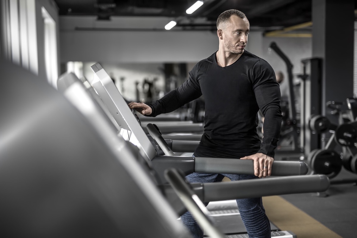 Handsome muscular man is preparing to run on the treadmill in the gym. He wears a black sweatshirt and blue pants. Guy looks backward. Shoot from the side. Horizontal.