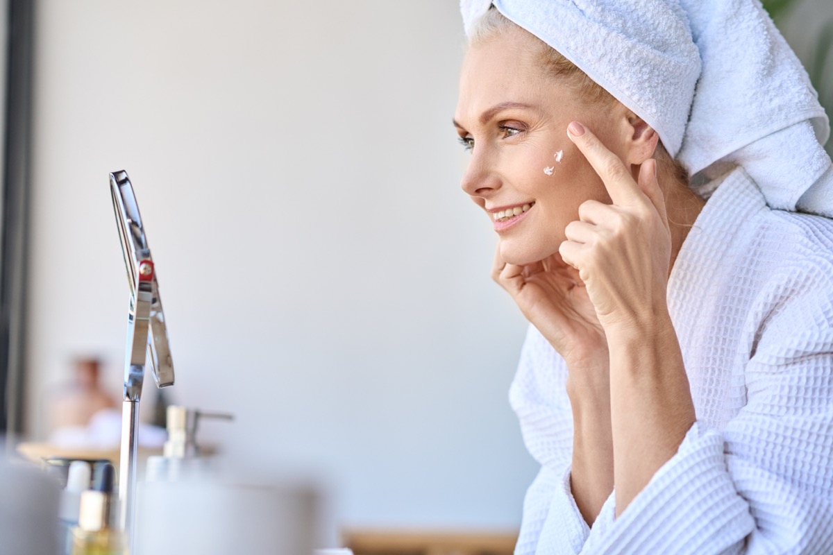 Gorgeous mid age older adult 50 years old blonde woman wears bathrobe in bathroom applying nourishing antiage face skin care cream treatment, looking at mirror doing daily morning beauty routine.