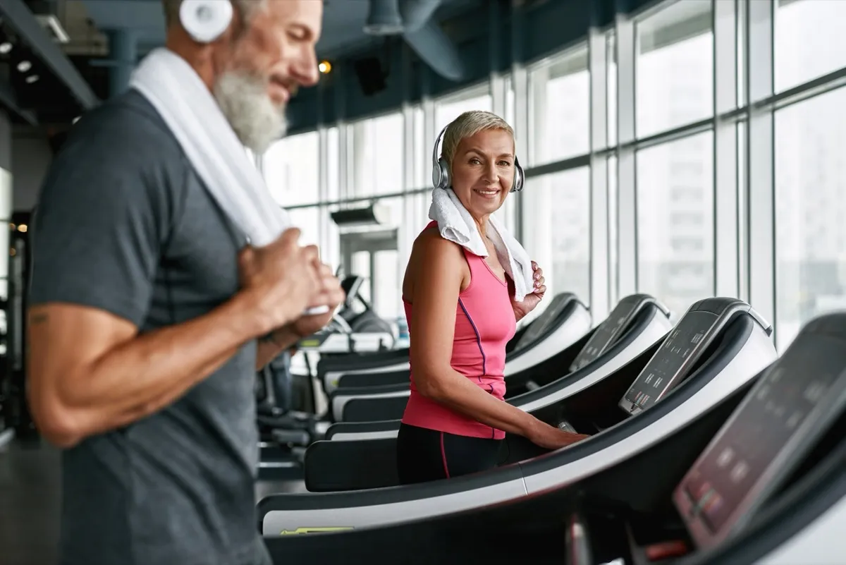 Portrait of good looking and fit elder woman working out on treadmill. Stylish senior woman looking younger than her age. Man with towel on shoulder on foreground, blured. Couple in gym.