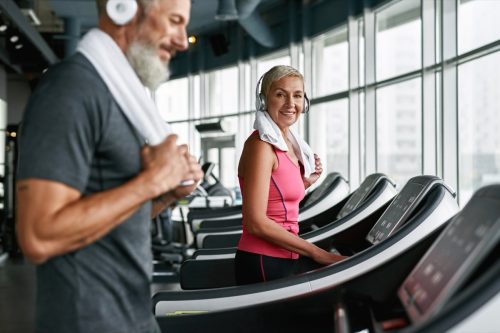 Senior man and woman on treadmills in the gym. The man in the foreground is looking down while the woman in the background smiles at him.