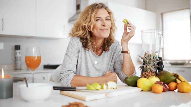 Portrait, fruit salad and apple with a senior woman in the kitchen of her home for health, diet or nutrition. Smile, food and cooking with a happy mature female pension eating healthy in the house