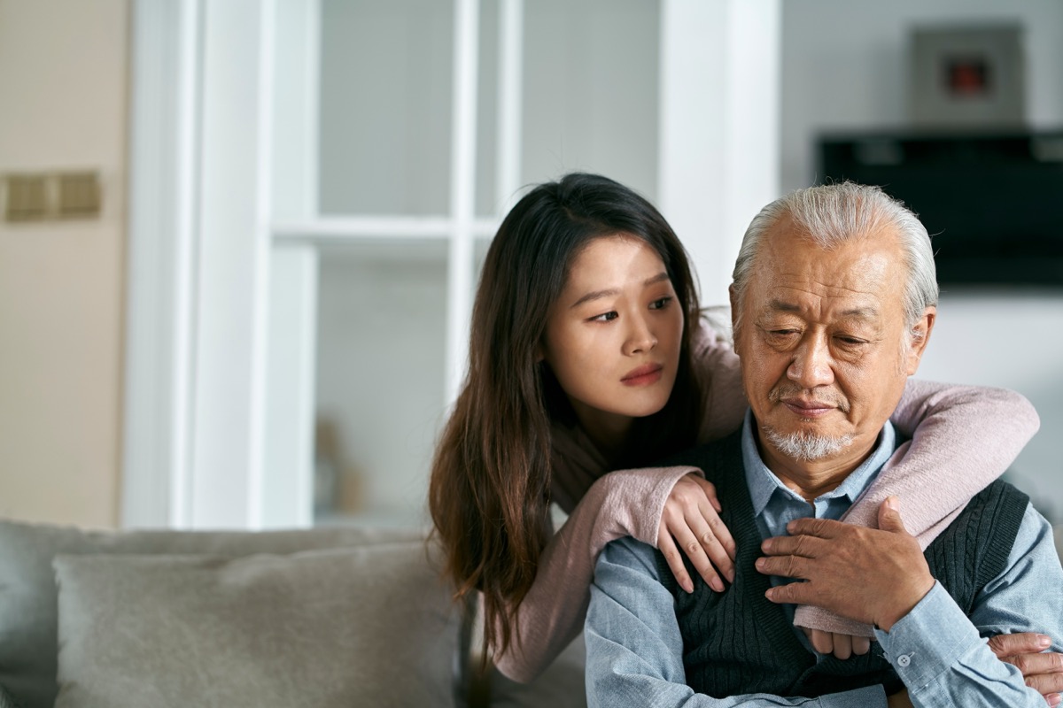 sad senior asian father sitting on couch in living room at home consoled by adult daughter