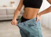 Great Weight Loss Result. Unrecognizable Fit Black Lady Showing Abdominal Muscles And Flat Belly Wearing Old Oversized Jeans After Successful Slimming Indoor. Cropped Shot