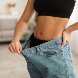 Great Weight Loss Result. Unrecognizable Fit Black Lady Showing Abdominal Muscles And Flat Belly Wearing Old Oversized Jeans After Successful Slimming Indoor. Cropped Shot