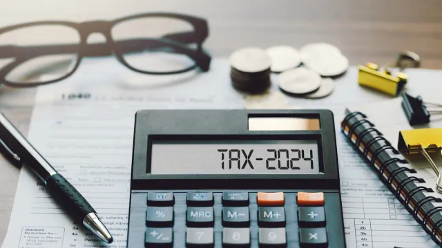 "Tax 2024" written out on a calculator among paperwork, glasses, pen, and coins.