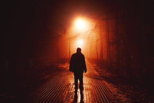 Sad man alone walking along the alley in night foggy park.