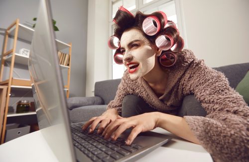 Mad woman in face mask and hair rollers sitting looking at laptop computer screen