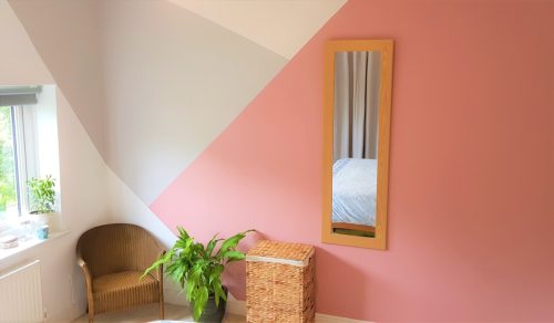 Bedroom image - geometric pattern painted on wall, grey cream and pink. Also contains a house plant, chair and laundry basket