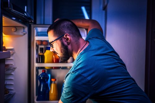 Young cheerful woman standing in front of the refrigerator at night, thinking about what to eat