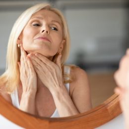 Middle aged woman touching skin on neck, looking at mirror in bedroom at home