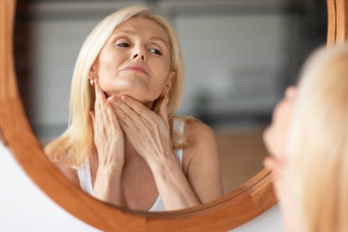 Middle aged woman touching skin on neck, looking at mirror in bedroom at home