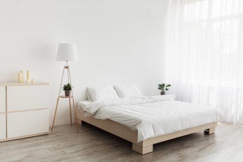 Simple modern design, ad, offer. Double bed with white pillows and soft blanket, lamp, furniture on wooden floor. White empty wall, big window with curtains in bedroom interior