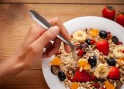 Top view close up of delicious, healthy homemade oats breakfast garnished with variation of mixed fruits toppings served in a white plate on wooden dining table and a hand picking a spoon full of it to eat.