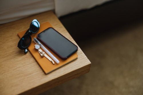 Close up shot of a bedside table in the hotel. There is a mobile phone, notebook, pen, watch and sunglasses.