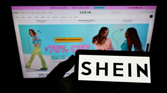 Person holding a smart phone with the Shein logo against a computer screen with the Shein homepage