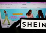 Person holding a smart phone with the Shein logo against a computer screen with the Shein homepage