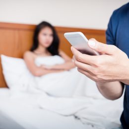 Cheating concept with unhappy couple – woman in bed while husband or boyfriend is texting