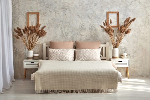 Large double bed with wooden headboard covered with wool plaid blanket and soft pillows standing in loft-style bedroom against gray cement wall with dry reed panicles in pots on night tables