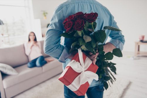 man holding a present and bouquet of roses behind his back to surprise his girlfriend on Valentine's Day