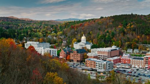 Aerial shot of the state capitol building in Montpelier, Vermont in Fall, flying over downtown buildings with a mountainside covered in colorful autumnal trees behind. 