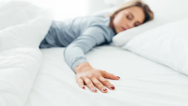 A woman in blue pajamas sleeping alone in her bed, with her arm draped over her partner's empty side.