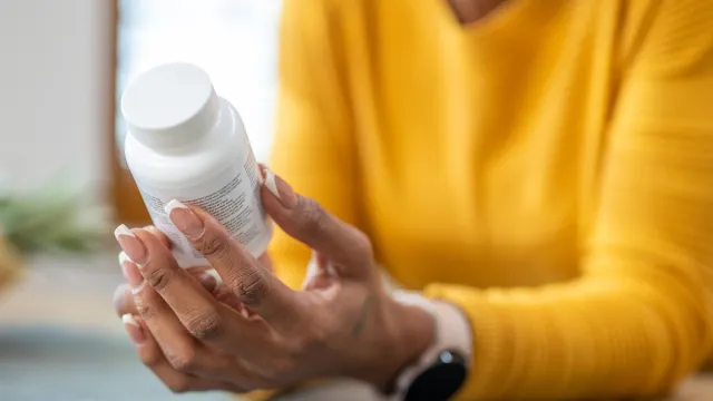 Close up of a woman in a yellow sweater holding a pill, vitamin, or supplement bottle, reading the ingredients