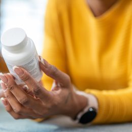 Close up of a woman in a yellow sweater holding a pill, vitamin, or supplement bottle, reading the ingredients