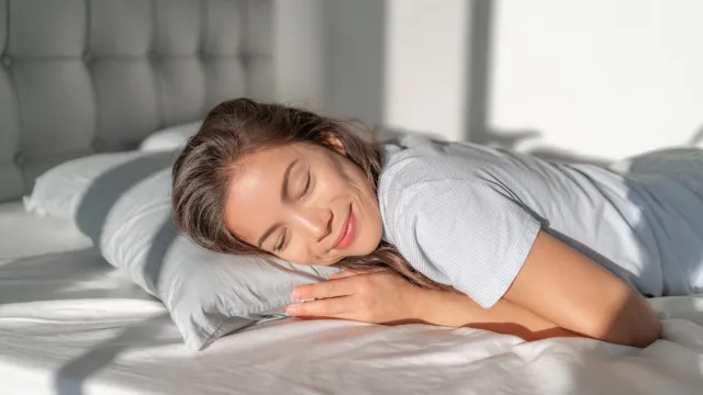 Young woman laying on her stomach on top of bed hugging her pillow and smiling.