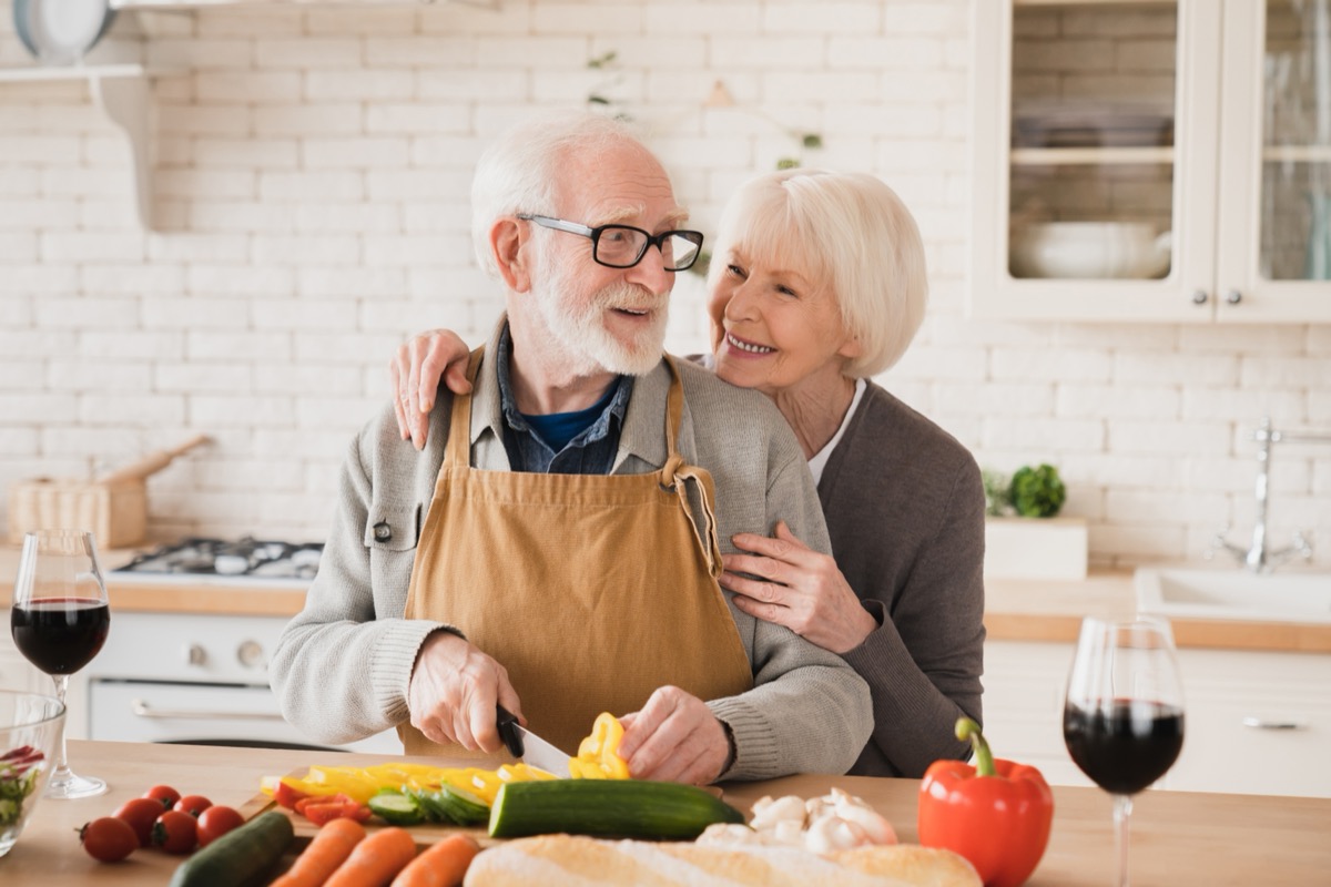 Senior couple chopping vegetables together in the kitchen while hugging and smiling