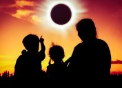Silhouette back view of family sitting and relaxing together. Boy point to solar eclipse on gold sky background. Happy family spending time together. Outdoor.