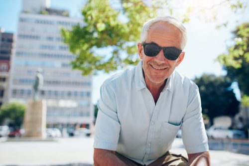 Portrait of happy senior man sitting outside in the city. He's wearing a blue short-sleeved button-down shirt and sunglasses.