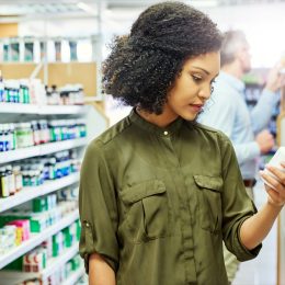 Pharmacy, health shop and woman with medicine for reading label, check product and choice in retail store. African female person looking at drugstore pills for ingredients, information and shopping
