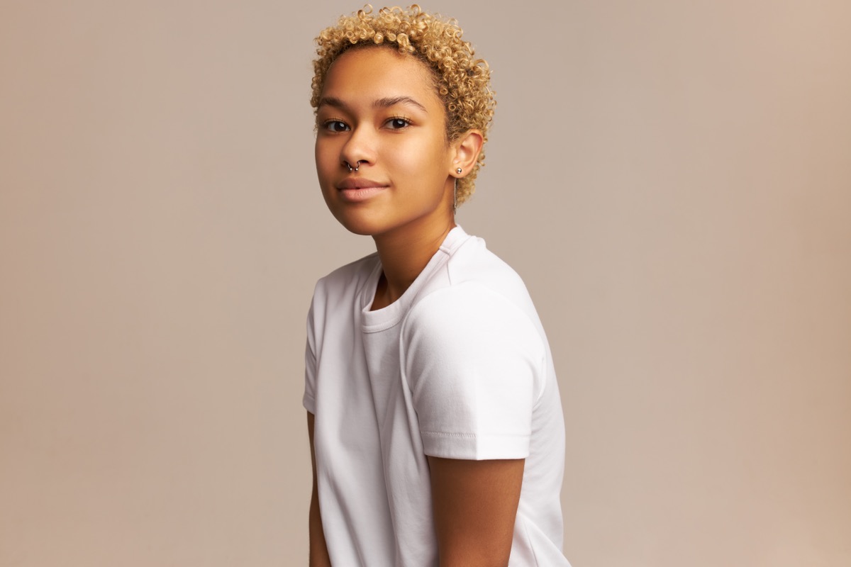Studio shot of attractive young lady of mixed-race posing with calm, peaceful face expression, gently smiling at camera, having trendy, natural look, wearing nose ring and short blonde haircut