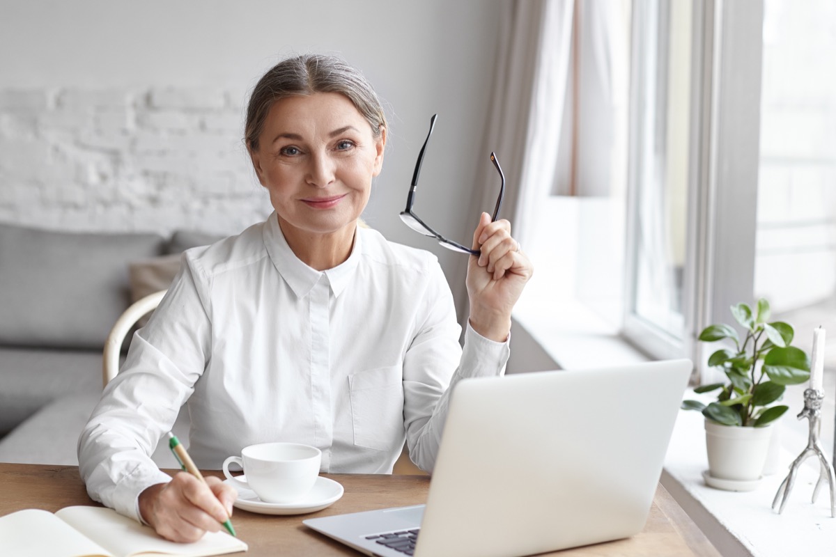 Elegant mature female psychologist working in modern office, holding eyeglasses and writing down in her diary, sitting at dews, using portable computer, smiling confidently, enjoying her work