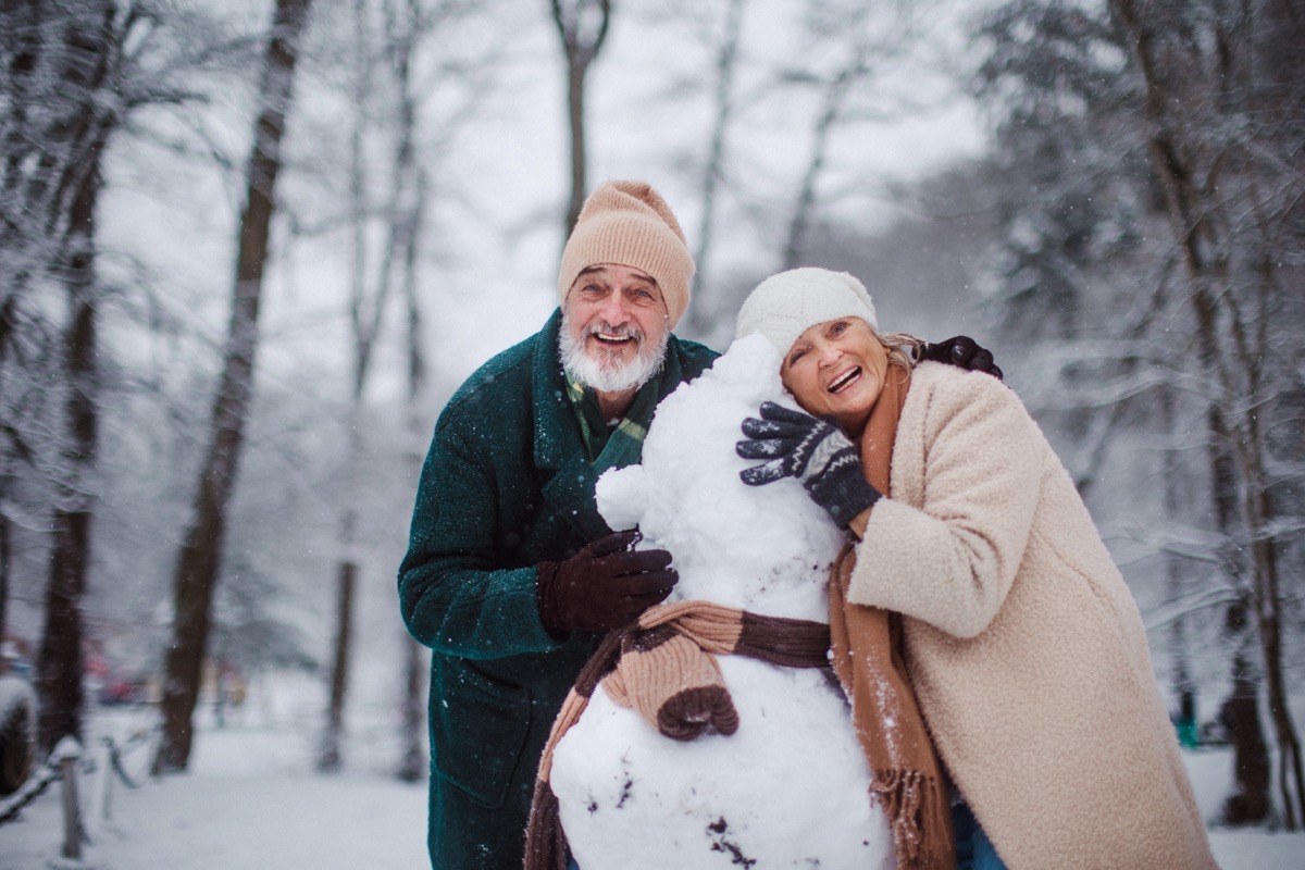 Elegant senior couple building snowman during cold winter snowy day.
