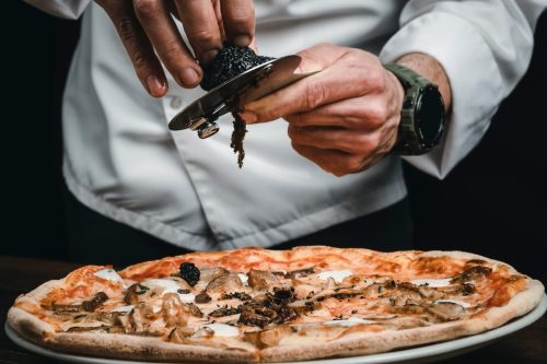 Close up of a man in a white chef's jacket shaving black truffle onto a mushroom pizza