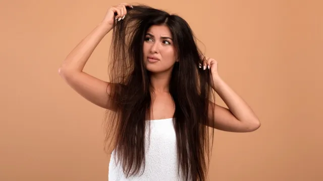 Frustrated armenian woman showing her damaged long locks on beige studio background. Young lady having bad hair day, upset over her messy hairdo. Hairdressing services concept