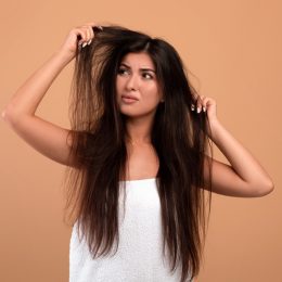 Frustrated armenian woman showing her damaged long locks on beige studio background. Young lady having bad hair day, upset over her messy hairdo. Hairdressing services concept