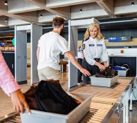 Two travelers putting their bags on the belt at airport security while a female TSA agent helps them.