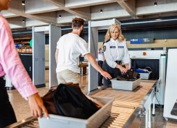 Two travelers putting their bags on the belt at airport security while a female TSA agent helps them.
