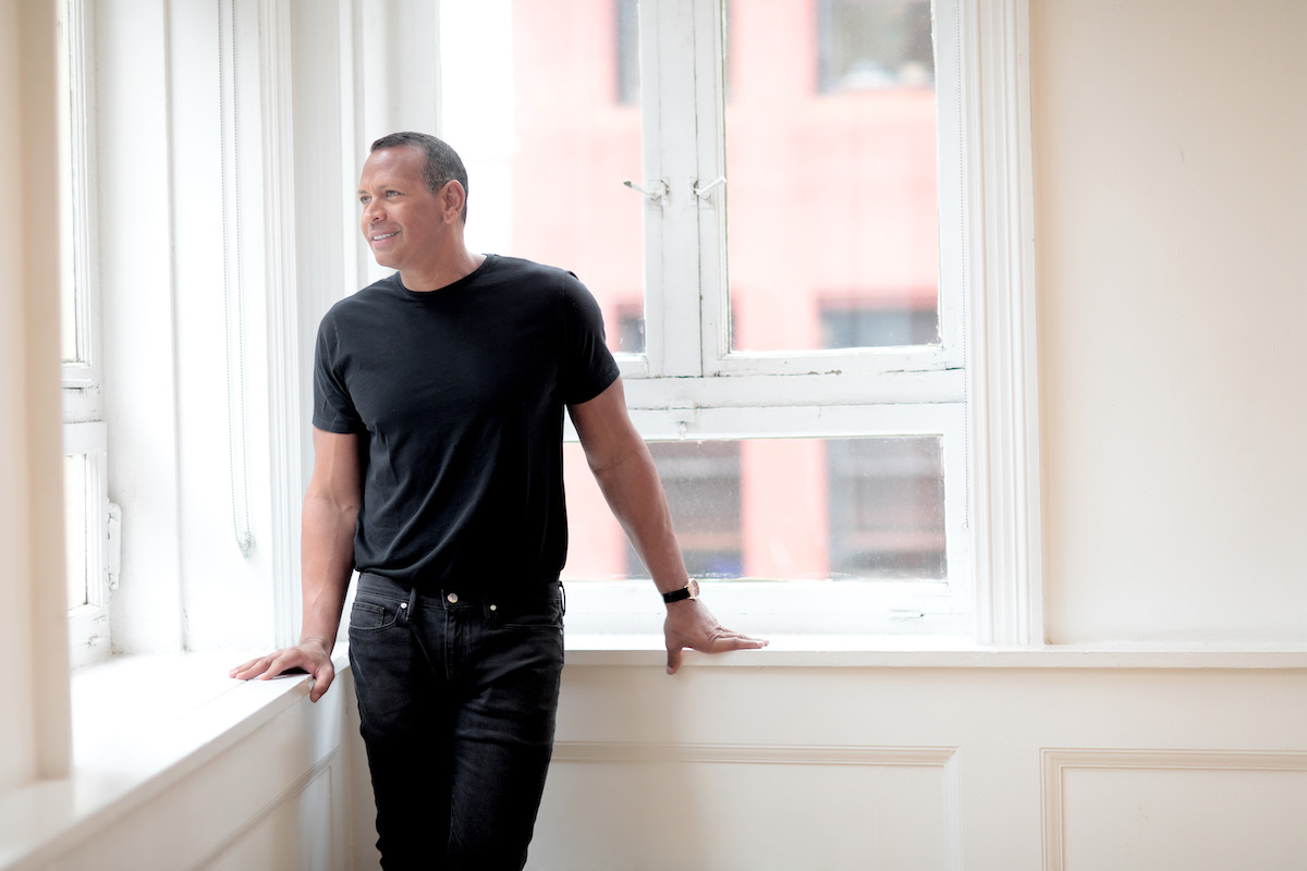 Alex Rodriguez wearing black jeans and a black t-shirt standing against white walls and large, city windows