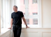 Alex Rodriguez wearing black jeans and a black t-shirt standing against white walls and large, city windows