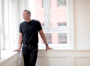 Alex Rodriguez wearing black jeans and a black t-shirt standing against white walls and large, city windows