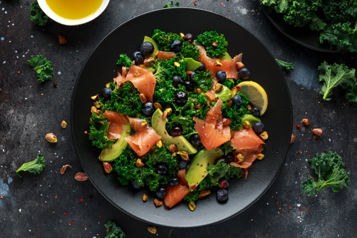 Salmon Kale super food Salad with avocado, pistachio nuts and blueberries on black plate.