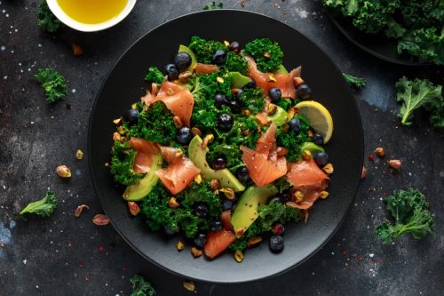 Salmon Kale super food Salad with avocado, pistachio nuts and blueberries on black plate.