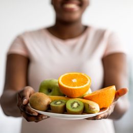 Blurred woman wearing a pale pink t-shirt holding out a plate of fruit, including oranges, kiwi, and banana.