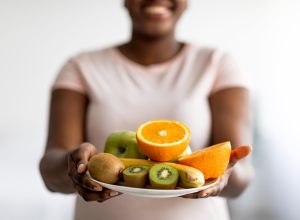 Blurred woman wearing a pale pink t-shirt holding out a plate of fruit, including oranges, kiwi, and banana.